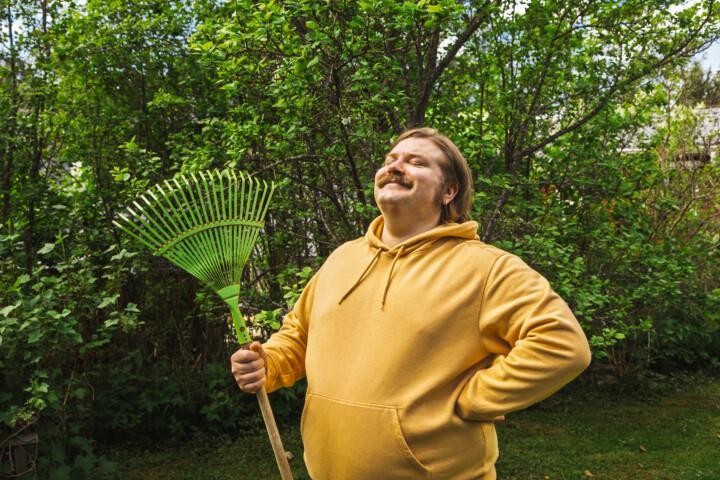 Henkilö seisoo vehreässä puutarhassa harava kädessä.